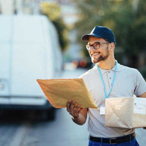 Young happy courier making a delivery in the city. Happy delivery man carrying packages while walking down the street.